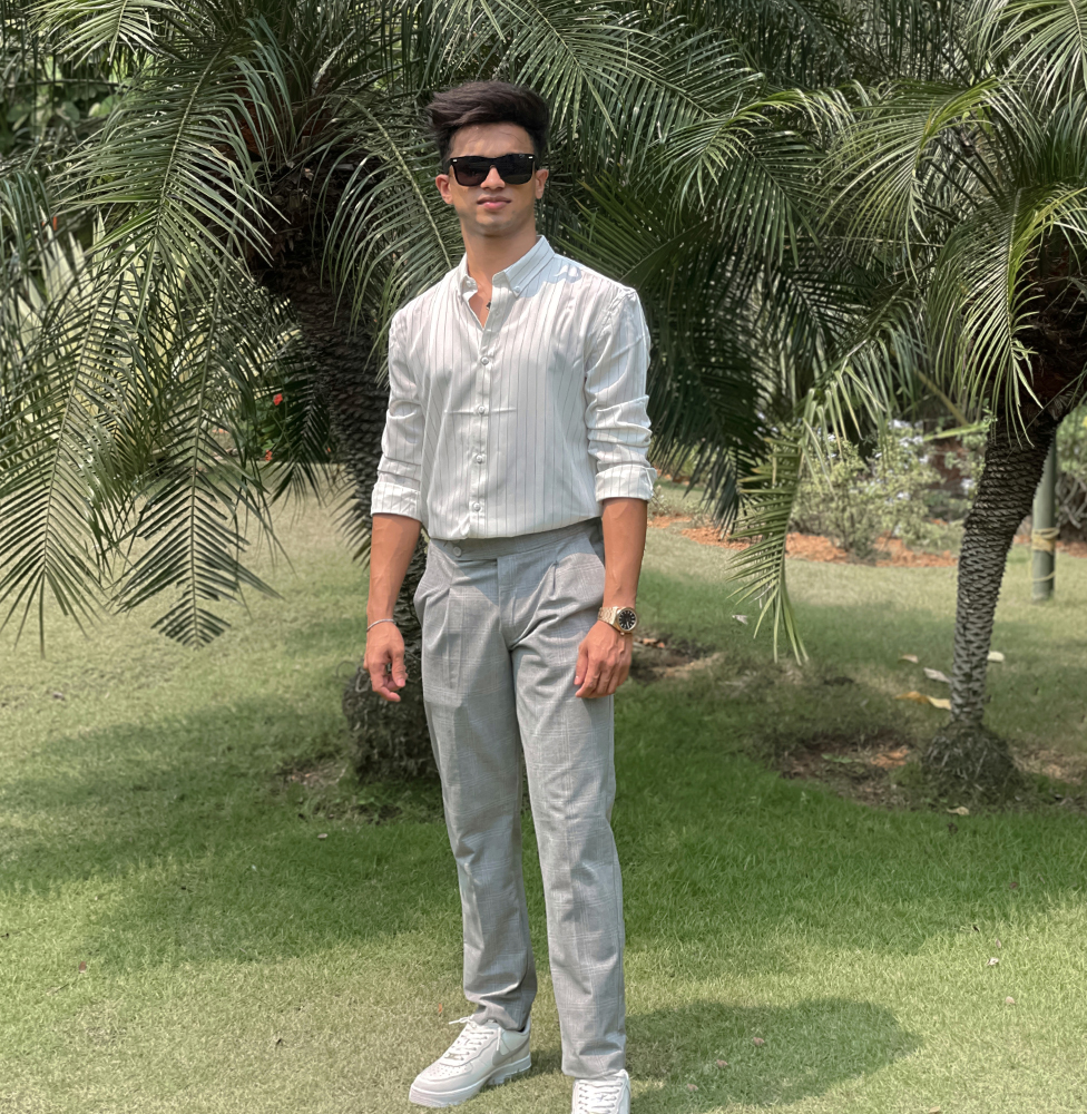 A young man with a watch and sunglasses stands confidently on a green lawn with palm trees in the background, wearing a white striped shirt, light gray pants, and white sneakers. Digital Marketer