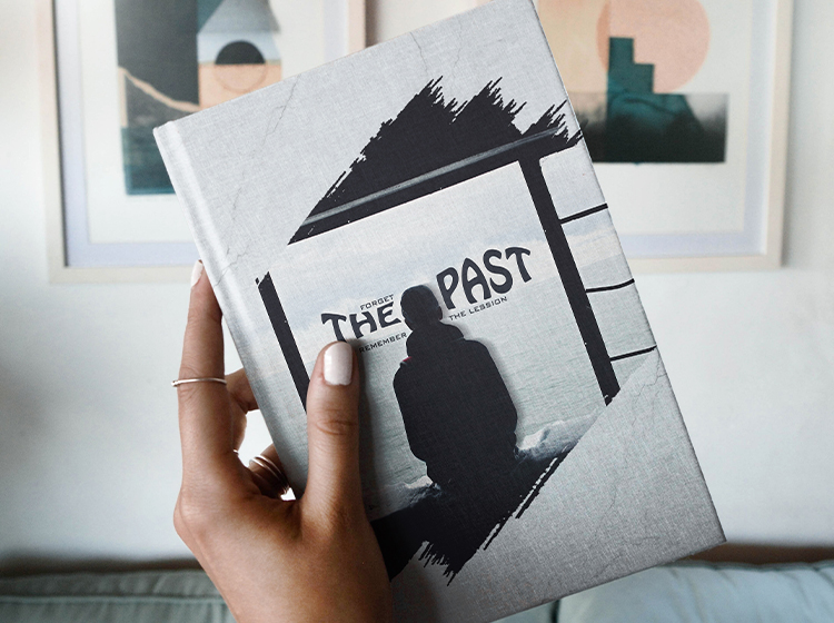 'Forget the Past, Remember the Lesson' and the silhouette of a person sitting are featured on the gray textured cover of a hardback book held in a hand. The background features a sofa and framed abstract wall art. Digital Marketer